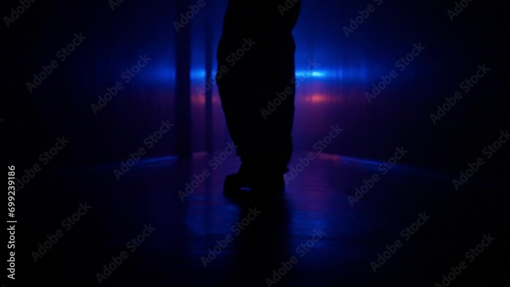 Portrait of silhouette in the hallway with neon light. Person in chemical protection suit feet close, walks cautiously in dark basement looks around, low angle.