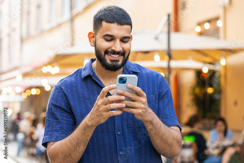Happy smiling bearded indian man using smartphone typing text messages browsing internet social media web app working chatting online outdoor. Guy tourist walking in urban city street. Town lifestyles