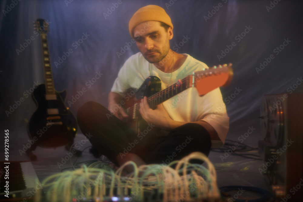 Obraz premium Blurry medium long shot of thoughtful adult male guitarist sitting legs crossed and playing electric guitar in makeshift studio