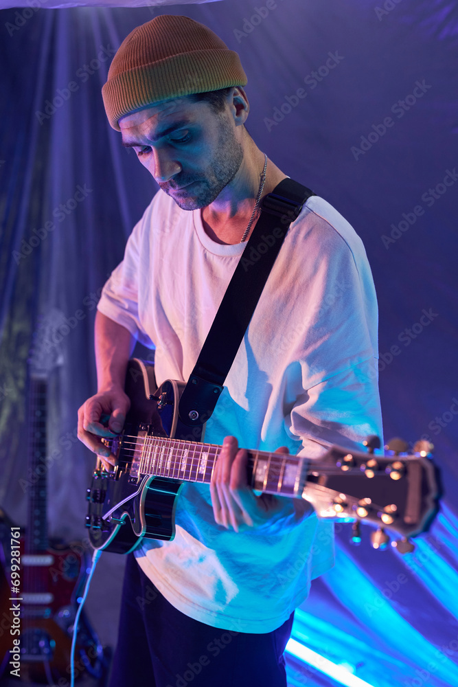 Obraz premium Vertical medium full shot of male band guitarist with stubble wearing beanie playing black semi-acoustic guitar on strap in blue neon light studio