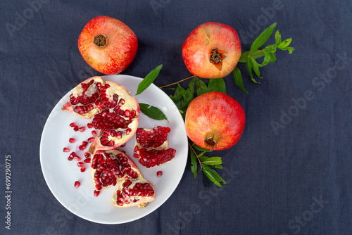 Three whole pomegranate fruits with a twig laying around a white plate with an opened fruit. Dark blue fabric table cloth. Top down view.