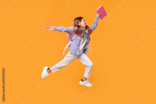Happy schoolgirl with book and backpack jumping on orange background