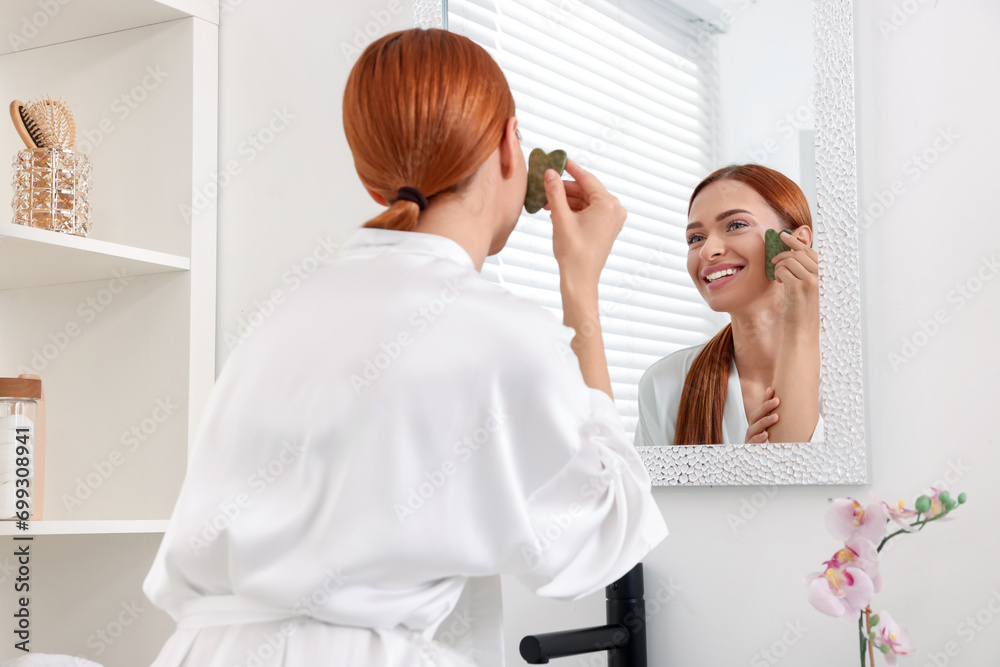 Young woman massaging her face with jade gua sha tool near mirror in bathroom Young woman massaging her face with jade gua sha tool near mirror in bathroom