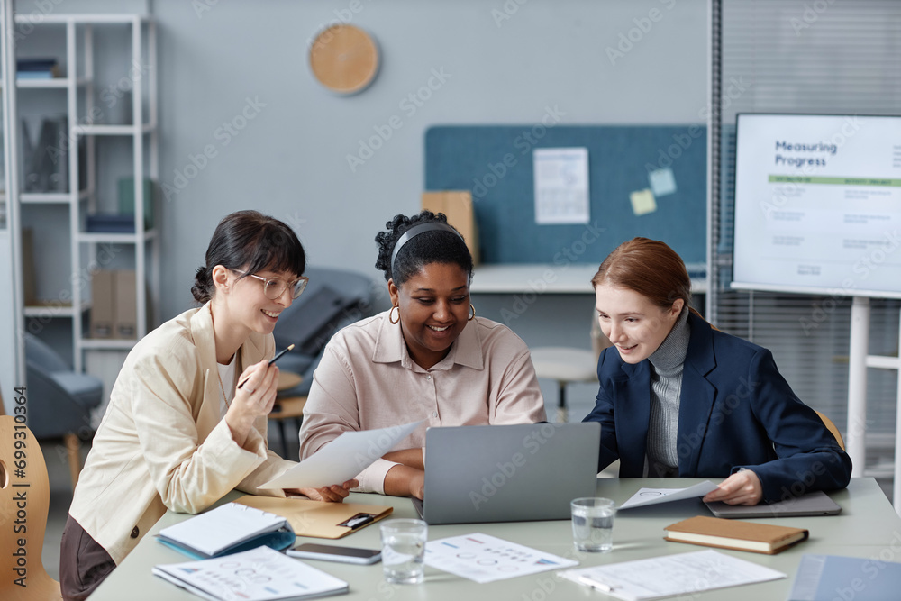 Medium shot of three cheerful diverse business women sitting at office desk looking at laptop