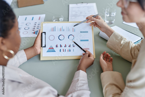 Over the shoulder shot of clipboard with multiple graphs, unrecognizable female business workers pointing at graphs on paper with pens