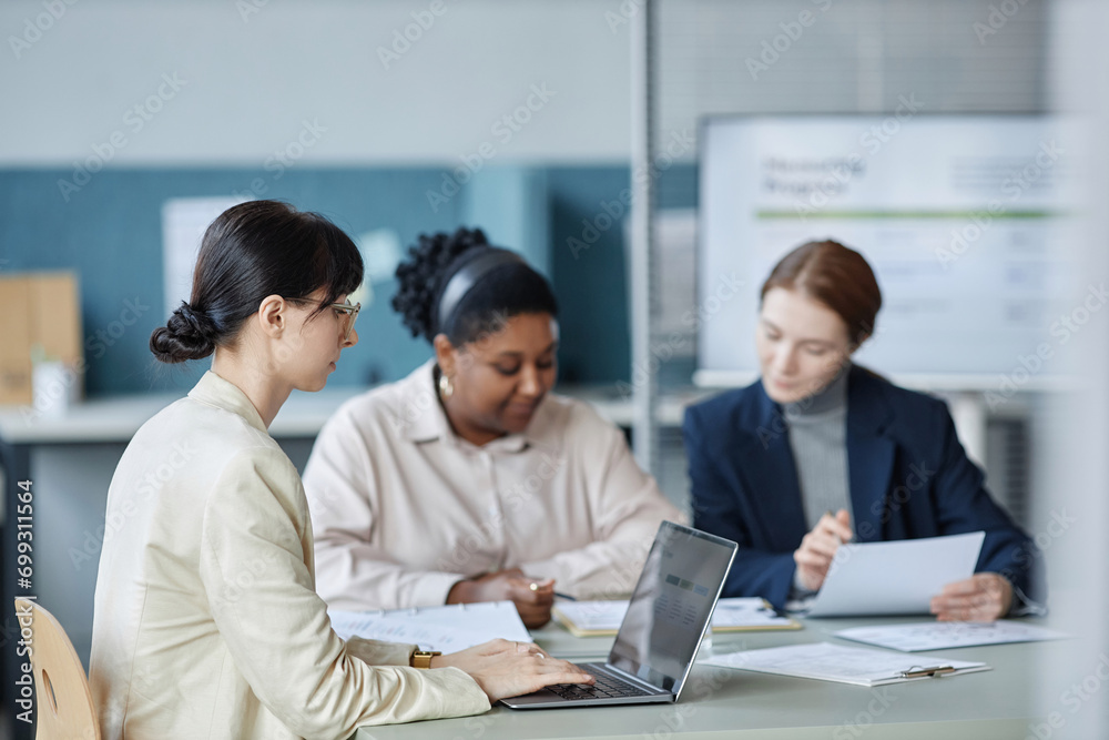 Side view of brunette business lady in beige suit working on laptop while sitting at office desk with female coworkers in blurred background