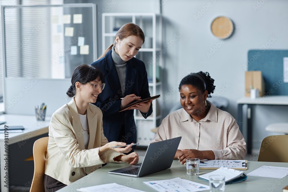 Medium shot of two cheerful diverse business women sitting at desk and looking at laptop while colleague in blue suit standing with tablet between them Medium shot of two cheerful diverse business women sitting at desk and looking at laptop while colleague in blue suit standing with tablet between them
