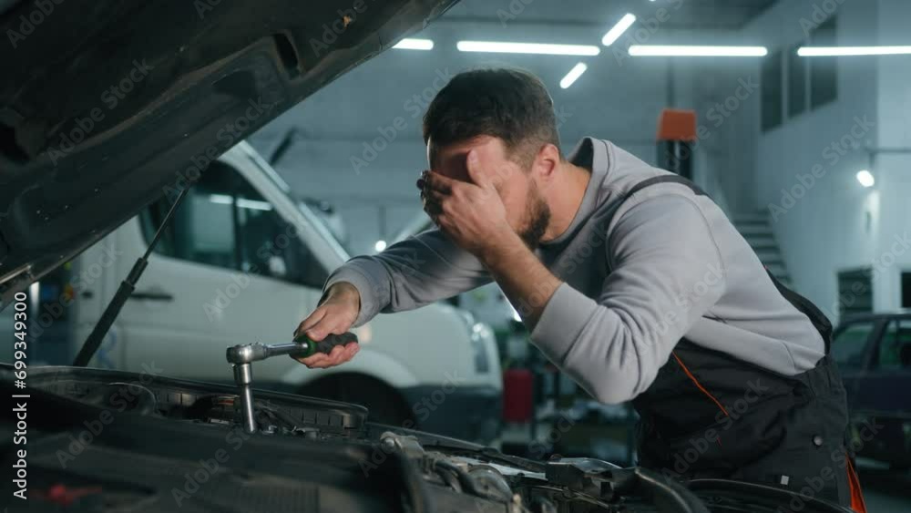Caucasian man tired exhausted auto mechanic overworking mending engine inside open car hood repairing vehicle with wrench tool at garage fixing broken automobile check maintenance in service station