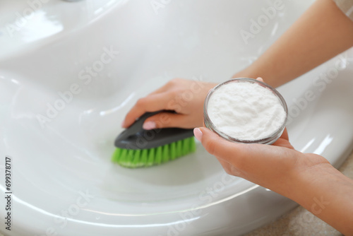 Woman cleaning white ceramic sink with brush and baking soda, closeup