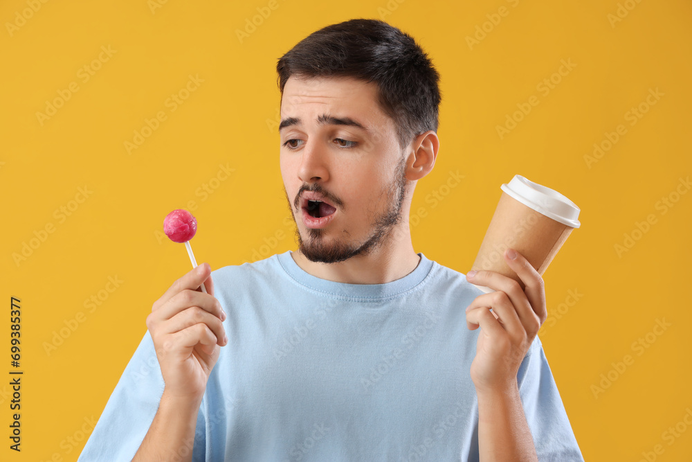 Handsome shocked young man with lollipop and cup of coffee on yellow background. Dental care concept Handsome shocked young man with lollipop and cup of coffee on yellow background. Dental care concept