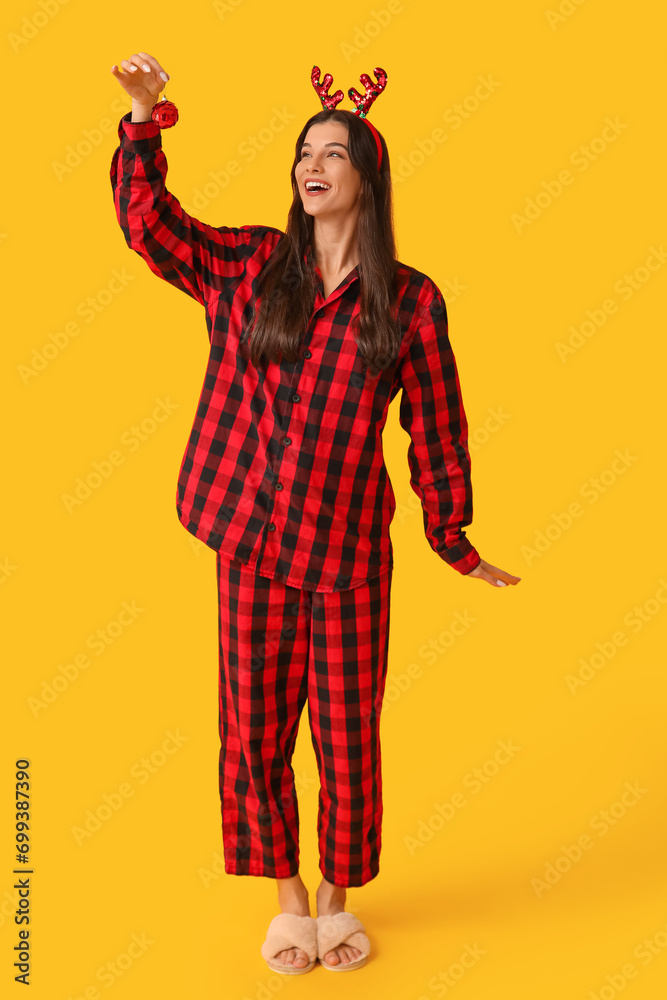 Beautiful young woman in checkered pajamas and reindeer horns with Christmas ball on yellow background