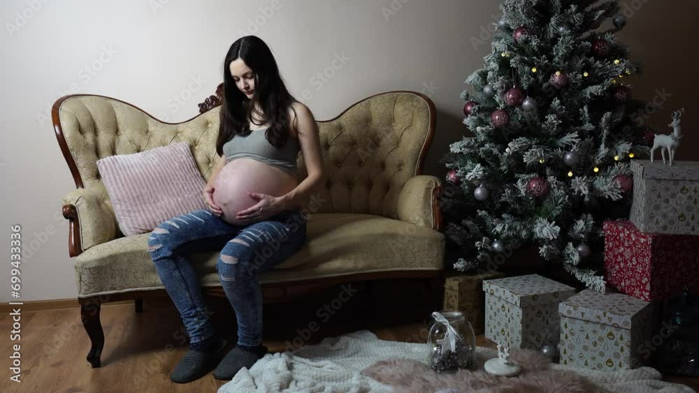 Pregnant woman sit near Christmas tree and softly touch big round belly Pregnant woman sit near Christmas tree and softly touch big round belly