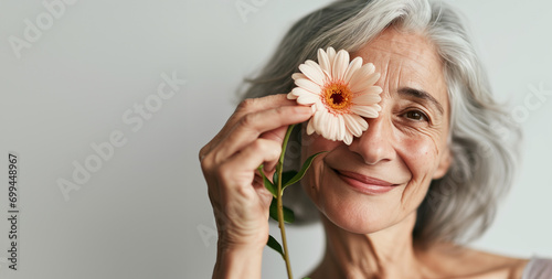 Photo portrait of senior 60 years old woman with fine lines and grey hair smiling looking at camera holding a flower next to her face, horizontal photo with copy space for banner