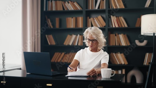 Senior woman freelancer works on laptop. Mature gray haired female in glasses working at home office writing notes typing on computer. Professional expert doing her job. Freelance online distant work. photo