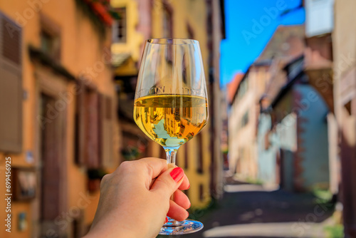 Woman s hand with a glass of white wine at an outdoor restaurant, blurred half timbered houses in Riquewihr, France, a village on Alsatian Wine Route photo