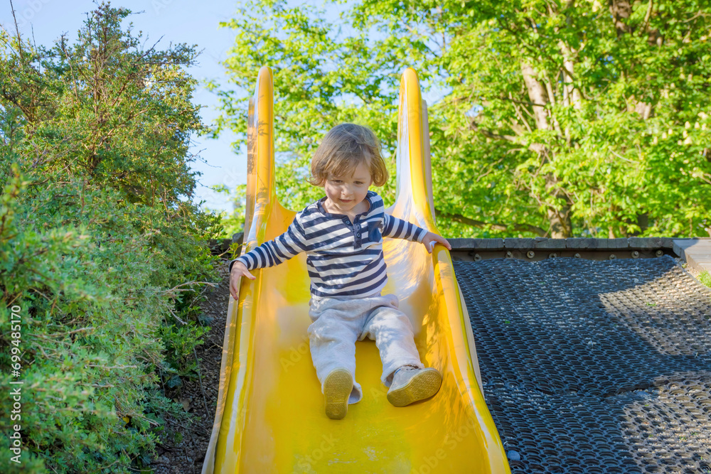 Happy cute little child boy having fun on a yellow slide outdoor in the park, sunny summer day in children playground. Selective focus