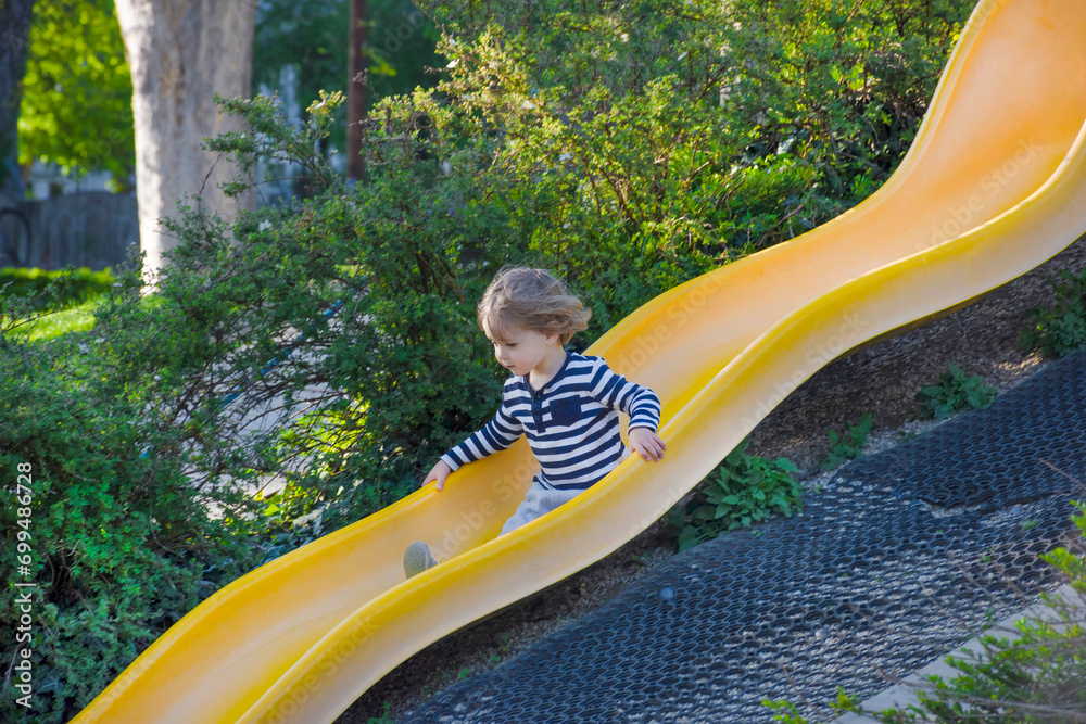 Happy cute little child boy having fun on a yellow slide outdoor in the park, sunny summer day in children playground. Selective focus