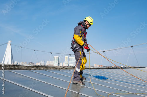 Worker on roof. Roofer carpenter roof construction. Male roofer replaces, repairs, installs roof of building. Guy standing in special protective suit with yellow helmet prepares ropes for belaying.
