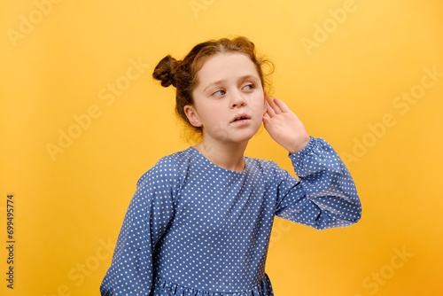 Portrait of excited caucasian preteen girl child overhearing tattles, listening with hand near ears to side, posing isolated over plain yellow color background wall in studio. Kids curiosity concept photo