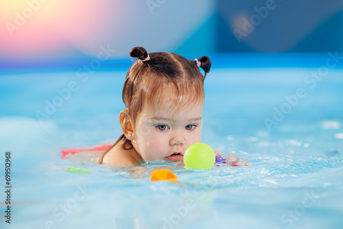 Happy baby girl swims and plays with balls in pool with trainer. Concept developing fine motor skills and health care of child