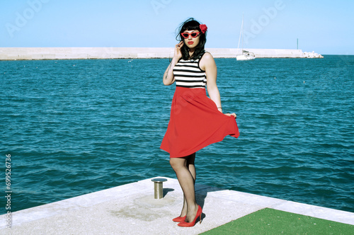 Port harbor pin-up: Red skirt, stripes, retro glasses. Red heels, intriguing secrets beneath: garter belt and sheer stockings. Revisited vintage elegance.