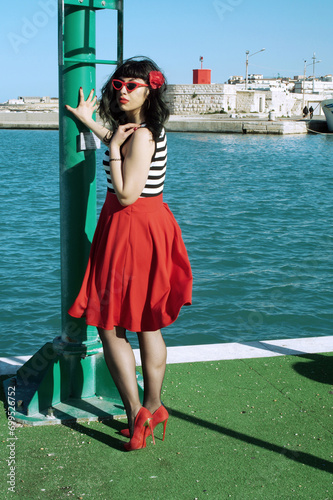 Port harbor pin-up: Red skirt, stripes, retro glasses. Red heels, intriguing secrets beneath: garter belt and sheer stockings. Revisited vintage elegance.