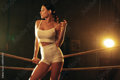 High fashion portrait of young beautiful sexy woman. Carefree model wearing white cycling shorts and tank top. Hot tanned female posing in boxing ring. Touches the rope. Cinematic. Smoke behind