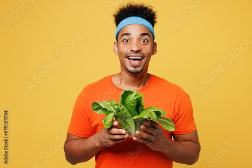 Young happy fitness trainer instructor sporty man sportsman wear orange t-shirt hold lettuce salad look camera spend time in home gym isolated on plain yellow background Workout sport fit abs concept