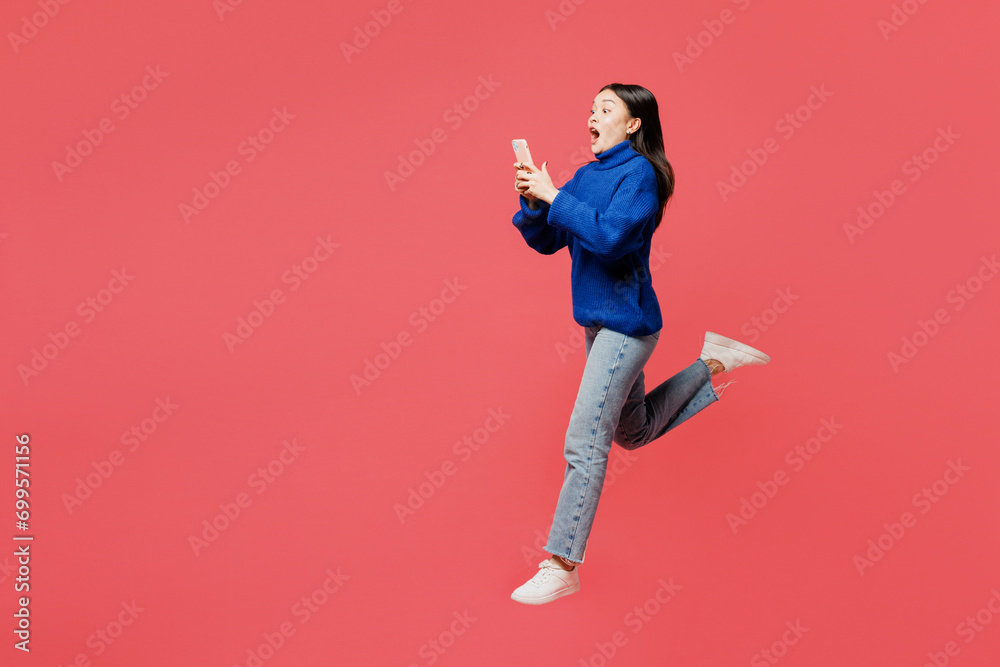 Full body side view young fun woman of Asian ethnicity she wear blue sweater casual clothes jump high hold in hand use mobile cell phone isolated on plain pastel light pink background studio portrait.
