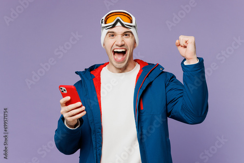 Skier happy man in warm blue windbreaker jacket ski goggles mask hat hold use mobile cell phone do winner gesture spend extreme weekend winter season in mountains isolated on plain purple background