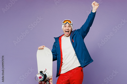 Happy man he wear warm blue windbreaker jacket ski goggles mask hat hold snowboard do winner gesture raise up hand spend extreme weekend winter season in mountains isolated on plain purple background. photo