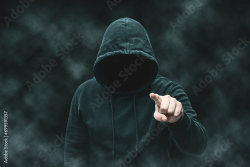 Mysterious faceless hooded anonymous criminal, silhouette of computer hacker, cyber terrorist or gangster on black background with smoke