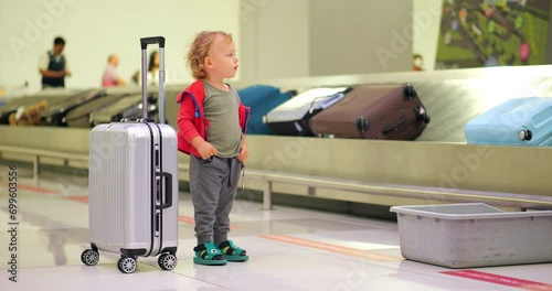 Young boy stand by trolley case at baggage claim hall area of large airport, slow motion full length portrait shot. Suitcases move on conveyor, seen blurred on background photo