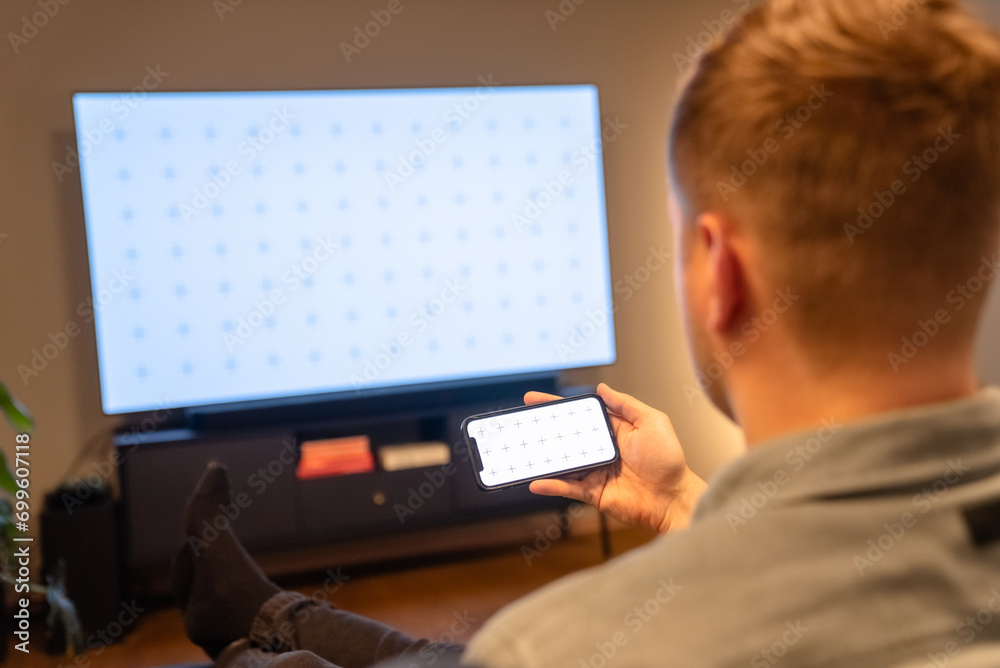 Man watching video on his phone while sitting on sofa, television in the background, watching movie Man watching video on his phone while sitting on sofa, television in the background, watching movie