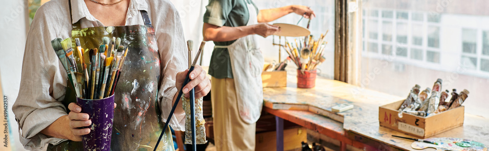 cropped view of mature female artists choosing paintbrushes in crafts workshop, horizontal banner