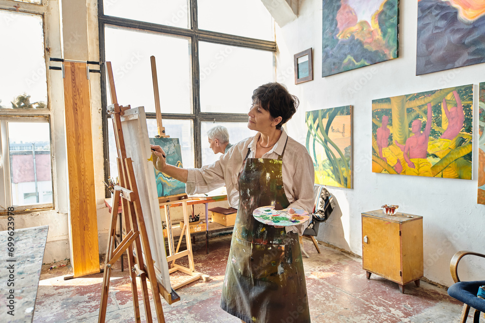 middle aged woman in apron painting on easel near female friend in modern spacious art workshop