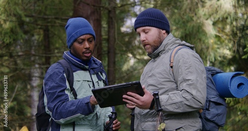 Two multiethnic tourists discuss trail way using tablet computer in forest. Outdoor enthusiasts with backpacks and trekking poles stop to rest during hike. Nature discovery and tourism. Slow motion. © Framestock
