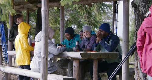 Group of diverse tourists resting in wooden gazebo after long walking trek in forest. Hiking buddies talk and laugh. Tourist friends during trip or expedition to mountains in autumn. Slow motion. © Framestock