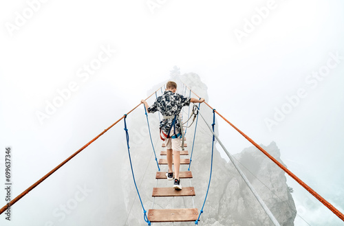 Courageous boy walks along rope bridge in mountain park on foggy day. Little tourist enjoys extreme attraction in cliffy valley. Sports activity photo