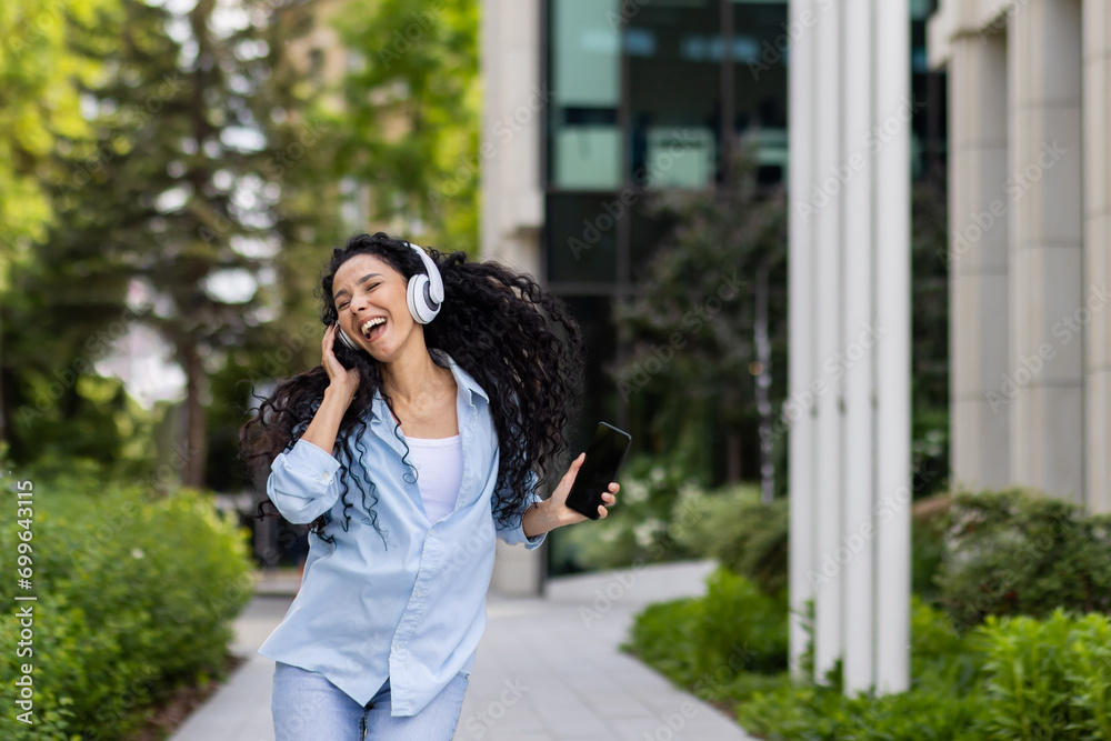 Young joyful beautiful woman dancing and singing in headphones while walking in the city, Hispanic woman with curly hair uses an application on the phone, to listen to music online. Young joyful beautiful woman dancing and singing in headphones while walking in the city, Hispanic woman with curly hair uses an application on the phone, to listen to music online.