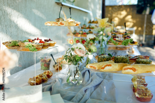 catering. Outdoor buffet table displays variety of appetizers on elevated glass platforms. Fresh flowers add aesthetic appeal to gourmet spread. Background reveals greenery, part of building structure photo