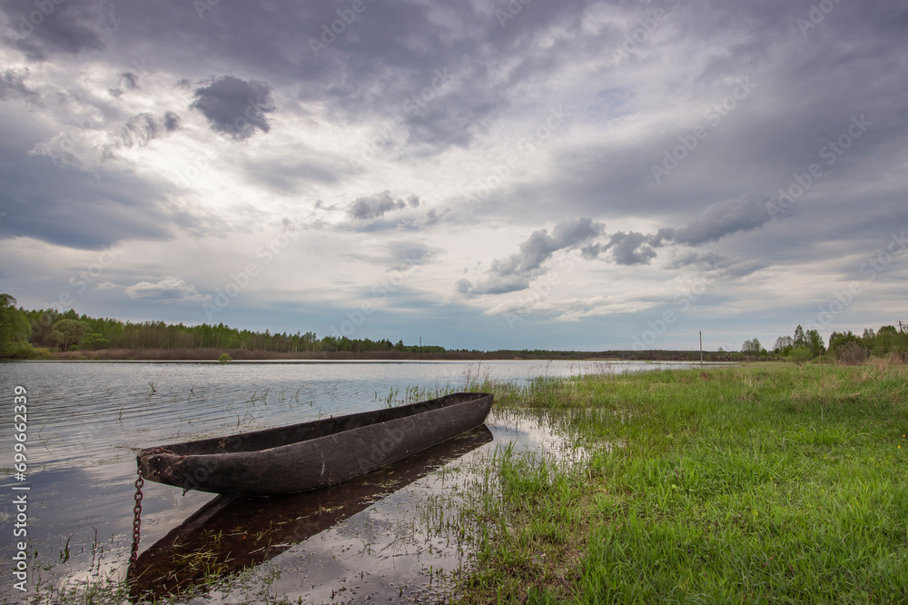 An ancient boat made from a whole tree trunk. Cloudy spring day by the river, boat in the foreground. An ancient boat made from a whole tree trunk. Cloudy spring day by the river, boat in the foreground.