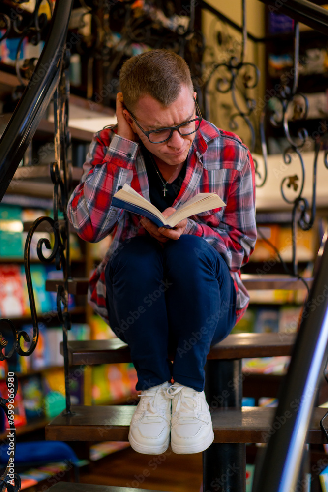 a man in glasses sits on the stairs in the library carefully reads a book and worries for its characters because of an unexpected plot line