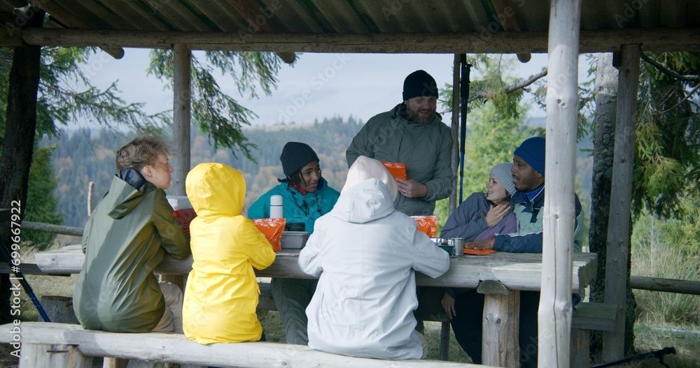 Diverse hiking buddies talk and prepare hiking food in pouches for dinner. Group of tourists rest in gazebo after walking trek in the forest. Hikers during trip or expedition to mountains on vacation. Diverse hiking buddies talk and prepare hiking food in pouches for dinner. Group of tourists rest in gazebo after walking trek in the forest. Hikers during trip or expedition to mountains on vacation.