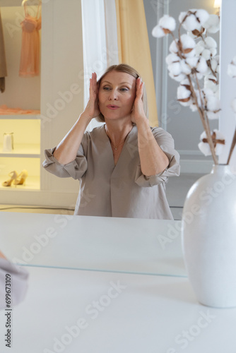 Skin care. Beautiful senior woman looking at herself at mirror doing face fitness - self face massage, sitting in front of mirror at home, making beauty routine procedures photo
