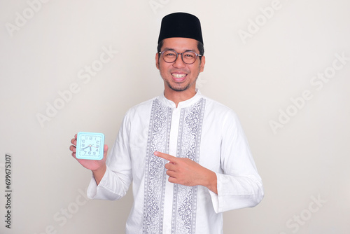 Moslem Asian man smiling at the camera while pointing to alarm clock that he hold photo