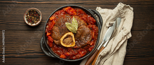 Traditional Italian dish Ossobuco all Milanese made with cut veal shank meat with vegetable tomato sauce served in black casserole pan top view on rustic brown wooden background.