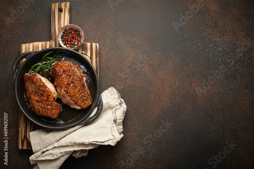 Two roasted duck breast fillets with crispy skin, with pepper and rosemary, top view in black cast iron pan with knife, dark brown concrete rustic background, space for text. photo
