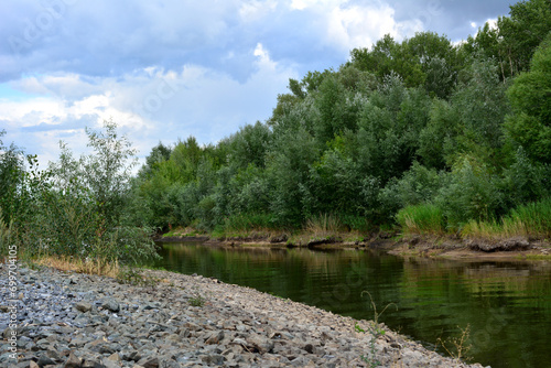 Fototapeta Naklejka Na Ścianę i Meble -  riverbank with gravel and calm water and green trees before rain copy space