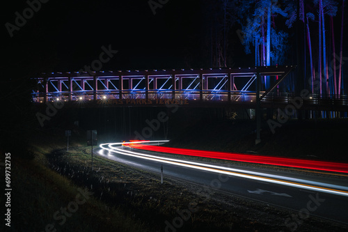 Colorful footbridge leading over a highway at night time. Light trails from car lighting up the road.  